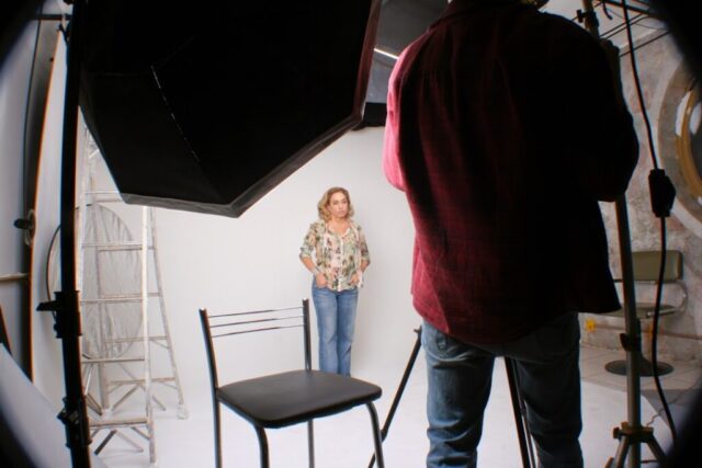 Cissa é uma mulher branca, de cabelos lisos, camisa florida e calça jeans. Ela olha para outra câmera durante gravação.