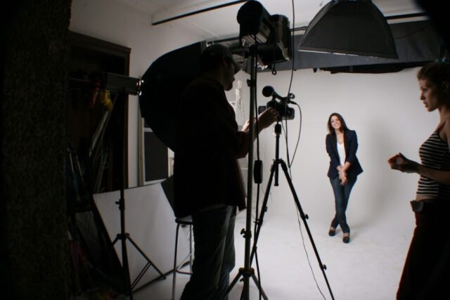 Carol é uma mulher branca, que veste camisa branca, blazer azul, calça jeans e calça sapatilha preta. Ela olha para outra câmera durante registro.